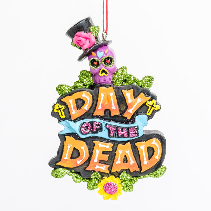 An ornament with "Day of the Dead" in orange with white bones framed with green glitter vines & a purple sugar skull on top 