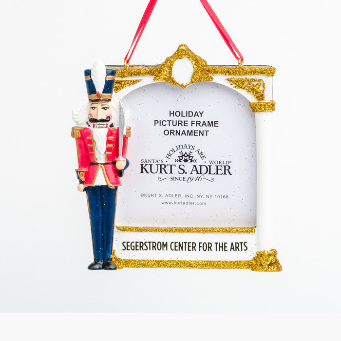 a white & gold glitter frame ornament with “Segerstrom Center for the Arts” at the bottom & a nutcracker in red & blue on the left